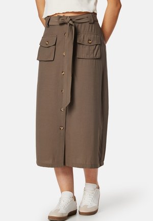 Khaki midi-rok met een gestrikt tailleband, twee voorzakken, knoopsluiting en een gladde textuur. Gepaarde met witte sneakers.
