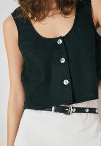 Un haut noir court en lin avec un col rond, orné de trois boutons transparents. Porté avec une ceinture noire à accents argentés sur une jupe blanche.