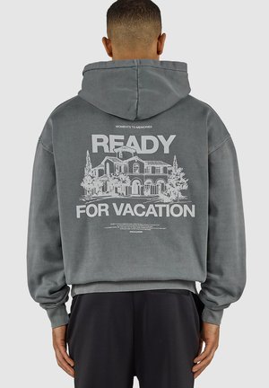 Mężczyzna w szarej bluzie z kapturem z napisem „READY FOR VACATION” i ilustracją domu na plecach, stojący na jednolitym tle.