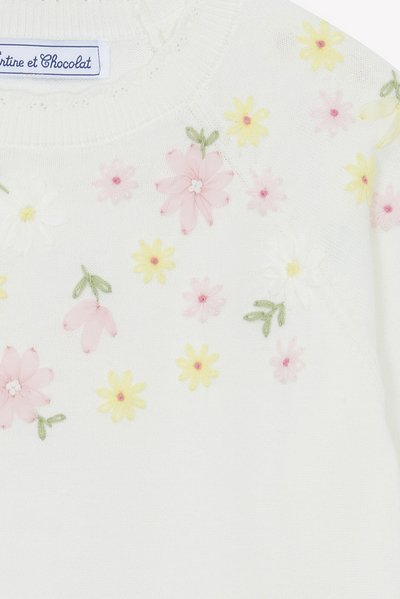 Pull en maille blanc avec broderie florale en rose, jaune et blanc. Présente un col rond et des coutures décoratives. Étiquette de la marque visible.
