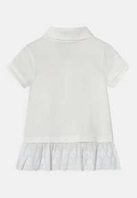 Vit pikétröja med krage och korta ärmar. Har en spetsdetalj vid fållen med blommönster. Slät textur genom hela plagg.