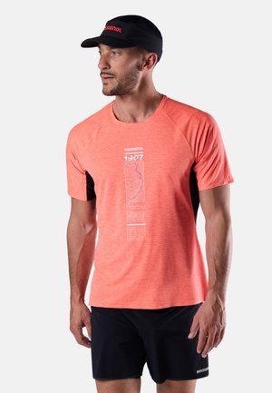 Homme portant un t-shirt de sport à manches courtes orange vif avec un motif, un short noir, regardant vers la gauche, et portant également une casquette visière noire.