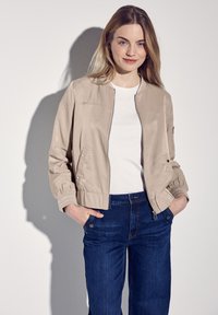 Světle béžová bomber bunda se zipem a elastickými manžetami, v kombinaci s bílým tričkem a tmavě modrými džíny. Hladký materiál, ležérní střih.