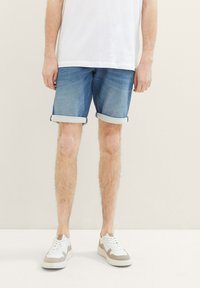 Hombre vestido con pantalones cortos de mezclilla azul con puños, zapatillas blancas y una camiseta blanca, de pie frente a un fondo beige liso.