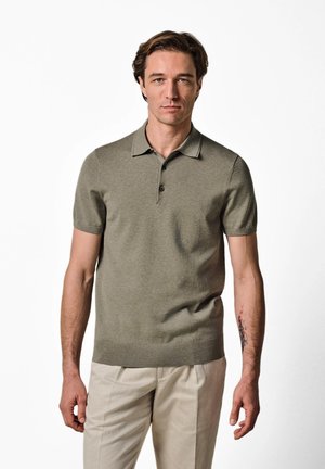 Hombre con cabello corto castaño, vestido con una camisa tipo polo verde de manga corta y pantalones beige, de pie frente a un fondo blanco liso.