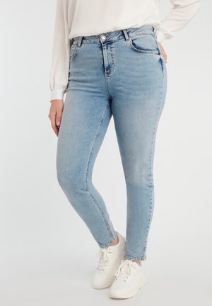 Person trägt hellblaue Skinny-Jeans, weißes Langarmshirt und weiße Sneakers und steht vor einem schlichten hellen Hintergrund.