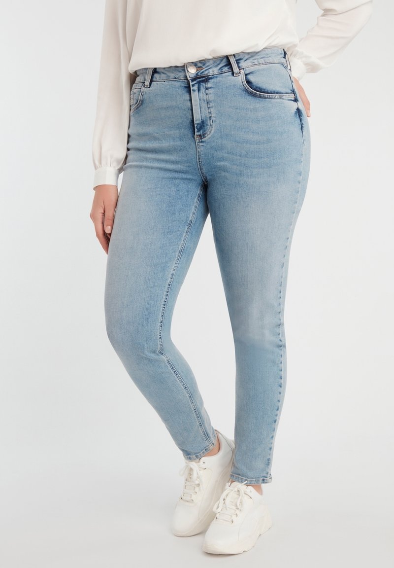 Personne portant un jean skinny bleu clair, une chemise blanche à manches longues et des baskets blanches, se tenant devant un fond clair uni.