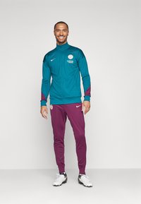 Teal takki, jossa on viininpunaiset yksityiskohdat ja valkoinen logo, yhdistettynä viininpunaisiin housuihin. Kevyt, urheilullinen kangas. Valkoiset kengät viimeistelevät asustuksen.