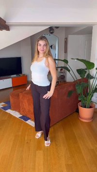 Young woman in white tank top and black pants standing in a living room with wooden floor, orange sofa, and large potted plant.