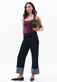 Cuerpo en púrpura profundo, pantalones de mezclilla oscuros de tiro alto con dobladillos en azul claro y costuras amarillas en contraste; sosteniendo un clutch negro.