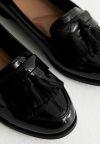 Zwarte gepatineerde leren loafers met een verhoogde kwastdetail, ronde neus en contrasterende stiksels langs de zool. Glad, glanzend textuur.