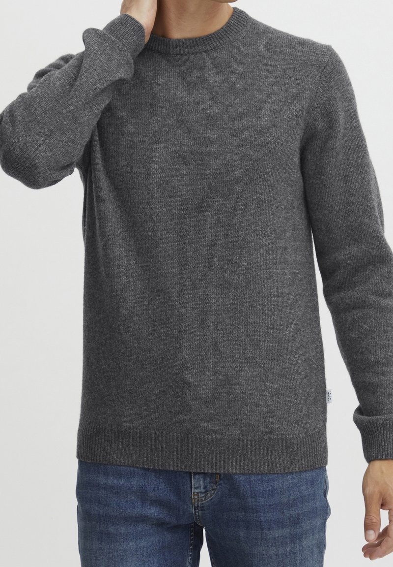Homme portant un pull en maille gris foncé uni avec des poignets et une bordure côtelés, associé à un jean bleu, sur un fond clair.