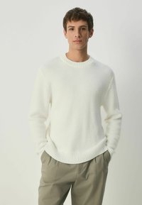 White knit sweater with ribbed collar and cuffs, featuring a textured pattern. Paired with light beige trousers, creating a casual look.