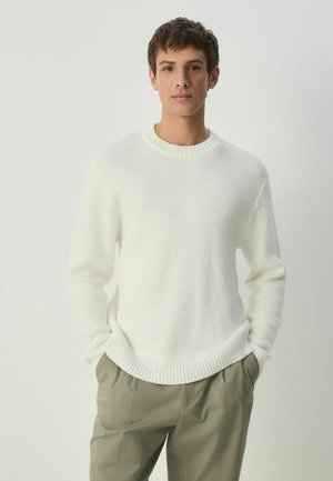 White knit sweater with ribbed collar and cuffs, featuring a textured pattern. Paired with light beige trousers, creating a casual look.