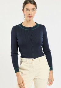 Cardigan bleu marine côtelé avec col et poignets à rayures vertes ; présente une fermeture à boutons, une coupe slim et des manches longues. Porté avec un pantalon de couleur claire.