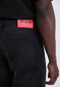 Jean en denim noir avec une étiquette de marque rouge indiquant "HUGO" à la ceinture, porté par une personne à la peau foncée avec des veines visibles sur les bras.