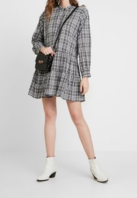 Grau kariertes Hemdkleid mit langen Ärmeln, gestuftem Saum und Kragen. Getragen mit einer schwarzen Umhängetasche und weißen Stiefeletten mit Absatz.