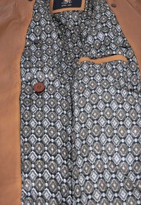 Braune Lederjacke mit einem gemusterten schwarz-weißen Futter, das geometrische Designs zeigt, zwei Knöpfen und einer kleinen Innentasche.