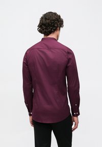 Bordowa koszula na guziki z długimi rękawami, zaokrąglonym dołem i subtelną fakturą. Rękawy ozdobione białymi guzikami na mankietach.