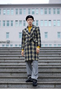 Abrigo de lana a cuadros en azul marino y amarillo con cintura anudada, combinado con un suéter de punto amarillo y pantalones de cuadros grises. Zapatillas negras completan el look.