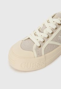 Sapatilha de lona bege com um upper texturizado, atacadores creme e uma sola de borracha grossa com um logo "GUESS" em relevo na lateral.