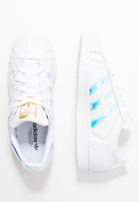 Bílé tenisky s duhově modrými a fialovými třemi pruhy. Mají texturovanou gumovou špičku a texturovanou bílou podrážku. Zlatý akcent s logem.