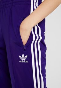 Lila Adidas joggingbyxor med vita ränder, ficka med dragkedja och logotyp. Tillverkade av mjukt material, med elastisk midja och åtsittande passform.