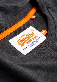 Tumma harmaa t-paita, jossa on pyöreä pääntie. Paidassa on suuri valkoinen etiketti, jossa on oranssia tekstiä "SuperDry" sekä lisäoranssia yksityiskohtaa kauluksessa.