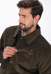 Sötétzöld kordbársony zakó nagy gallérral, gombos záródással és hímzett horgonylogóval. Finom szöveges részleteket tartalmaz az anyagon.