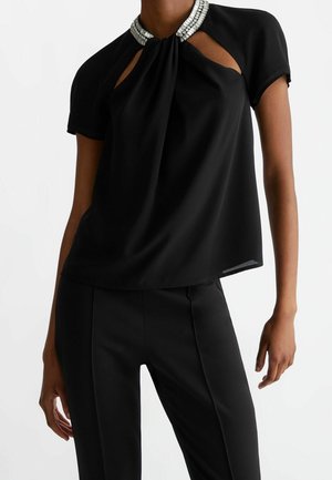 Femme portant un haut noir à manches courtes avec un col orné de strass et des découpes en forme de trou de serrure, associé à un pantalon noir cintré.