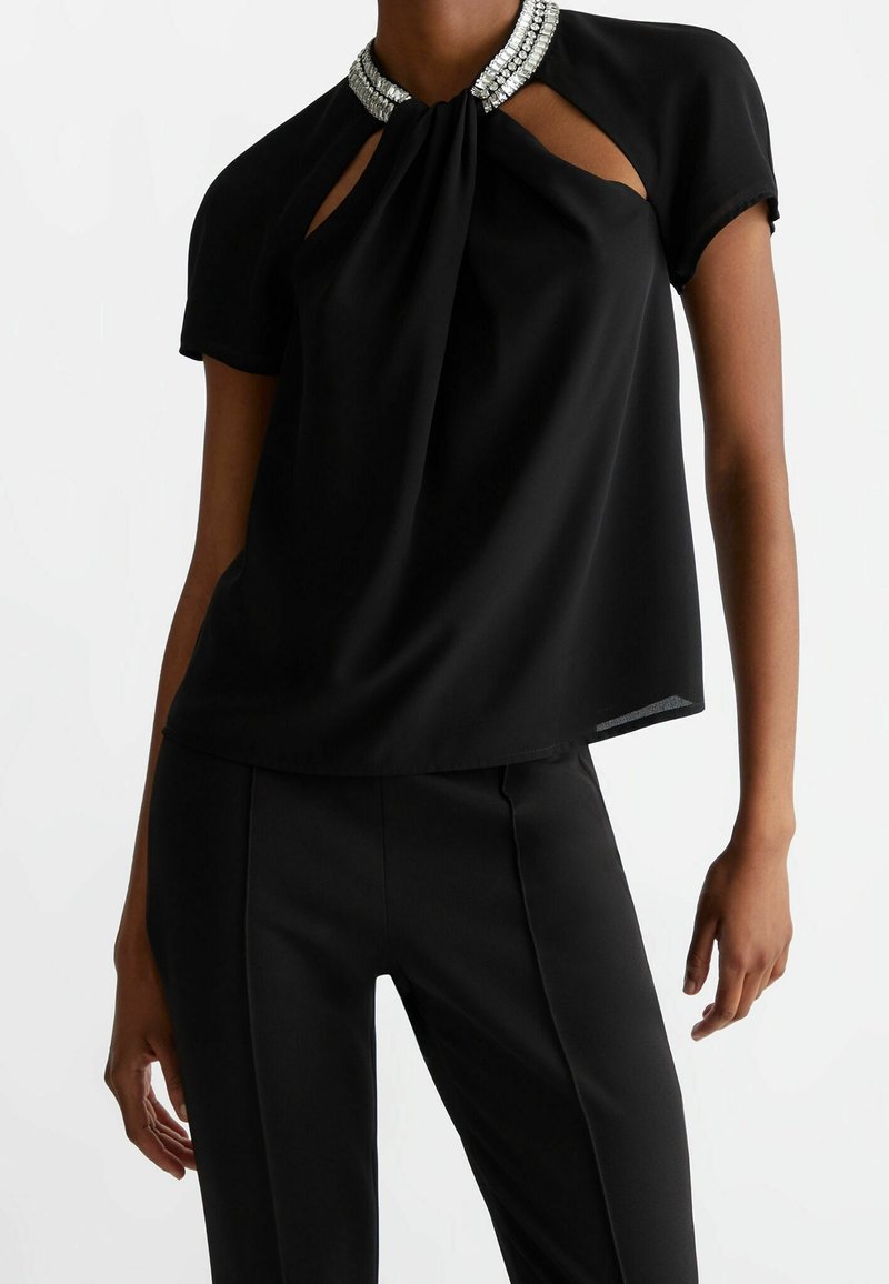 Femme portant un haut noir à manches courtes avec un col orné de strass et des découpes en forme de trou de serrure, associé à un pantalon noir cintré.