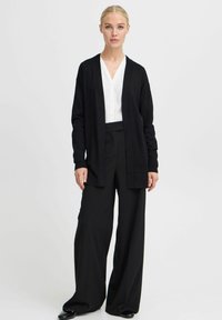 Cardigan long noir en tissu doux, avec un design à devant ouvert. Porté sur une chemise blanche et un pantalon large noir, créant un look monochromatique.