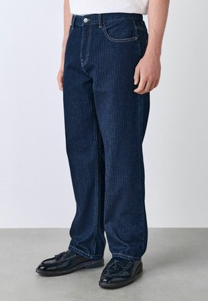 Dunkelblaue Jeans aus Denim mit geradem Bein, mit einem Knopfverschluss, fünf Taschen und dezenten vertikalen Streifen. Kombiniert mit schwarzen Schuhen.