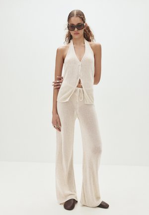 Top halter in maglia color beige chiaro con chiusura a bottone e pantaloni abbinati svasati. Texture morbida, vestibilità ampia e design a gamba larga. Scarpe slip-on marroni.