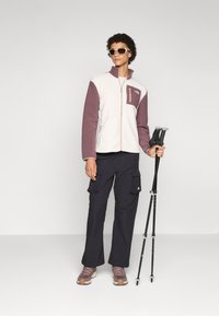 Fleece jack met crèmekleurige en mauvepanelen, met een ritsborstzak; gecombineerd met zwarte cargobroek en bruine wandelschoenen, terwijl hij trekkingstokken vasthoudt.