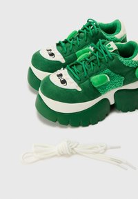 Sneakers in suede verde con suola bianca chunky, caratterizzate da occhi di cartone sulla punta e un accento peloso verde. Comprendono lacci bianchi.