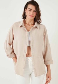 Chemise beige clair à manches longues et boutons, en tissu texturé. Comprend un col, une poche poitrine et des poignets retroussés. Associée à un débardeur blanc.