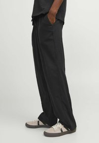 Henkilö, jolla on päällään löysät mustat housut ja beiget lenkkarit, joissa on mustat raidat, seisoo valkoista taustaa vasten, toinen käsi taskussa.