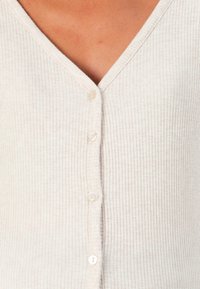 Ljust beige ribbat topp med V-ringning och sex små, vita knappar framtill. Materialet verkar mjukt med ett strukturerat mönster.