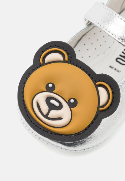 Zapato metálico plateado con una aplicación de cara de oso negro y marrón. El oso presenta acentos blancos para los ojos y la nariz, creando un diseño juguetón.