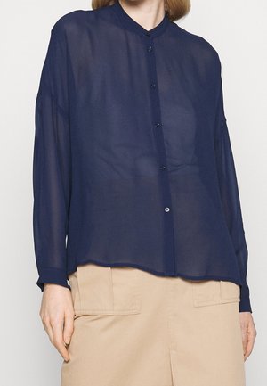 Femme portant une chemise boutonnée à manches longues, en voile bleu marine, associée à une jupe beige avec de grandes poches avant.