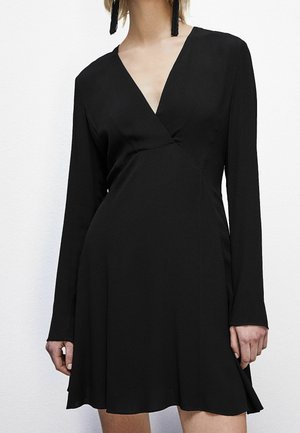 Femme portant une robe noire à manches longues avec un col en V plongeant et un détail asymétrique à la taille, assortie à de longues boucles d'oreilles noires à pampilles.