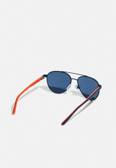 Óculos de sol estilo aviador com lentes azul-escuro, armação de metal preto e hastes de plástico vermelhas com uma faixa de destaque azul.