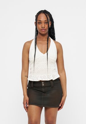 Woman with long braided hair, wearing a white sleeveless eyelet top and a black denim mini skirt, standing against a plain background.