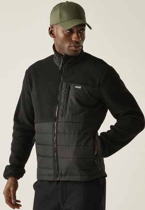 Uomo con giacca in pile nera con sezioni trapuntate e tasche con cerniera, e cappellino da baseball verde oliva, che guarda di lato.