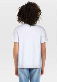 Biała koszulka z krótkim rękawem i okrągłym dekoltem. Wykonana z gładkiej bawełny. Tył jest gładki, bez logo ani grafik. Widoczne dżinsy poniżej.