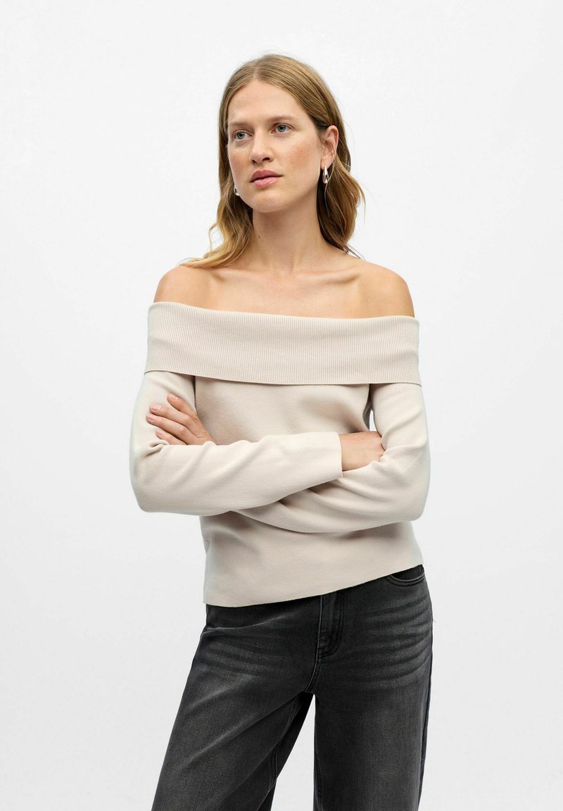 Svetlo bež pulover z odprtimi rameni, z rebrastim ovratnikom, dolgimi rokavi in prilegajočo silhueto. Usklajen s temno sivo jeans hlačami.