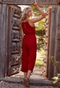 Combinaison rouge avec une taille ajustée et des bretelles, dotée d'un dos en keyhole et de jambes larges. Portée avec des sandales marron, devant des cadres de porte en bois.