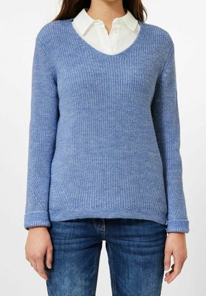 Maglione azzurro lavorato a maglia con scollo a V, texture a coste e polsini risvoltati, abbinato a una camicia bianca con colletto sotto.