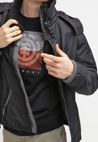 Czarna kurtka z teksturowanymi ramionami, zapięciem na zamek i szarymi akcentami, noszona na czarnym T-shircie z geometrycznym czerwonym wzorem graficznym.